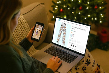 Woman shopping personalized polyester pajama pants printed with her dog’s photo on Duconspace website, holding her pet’s picture on phone beside Christmas tree