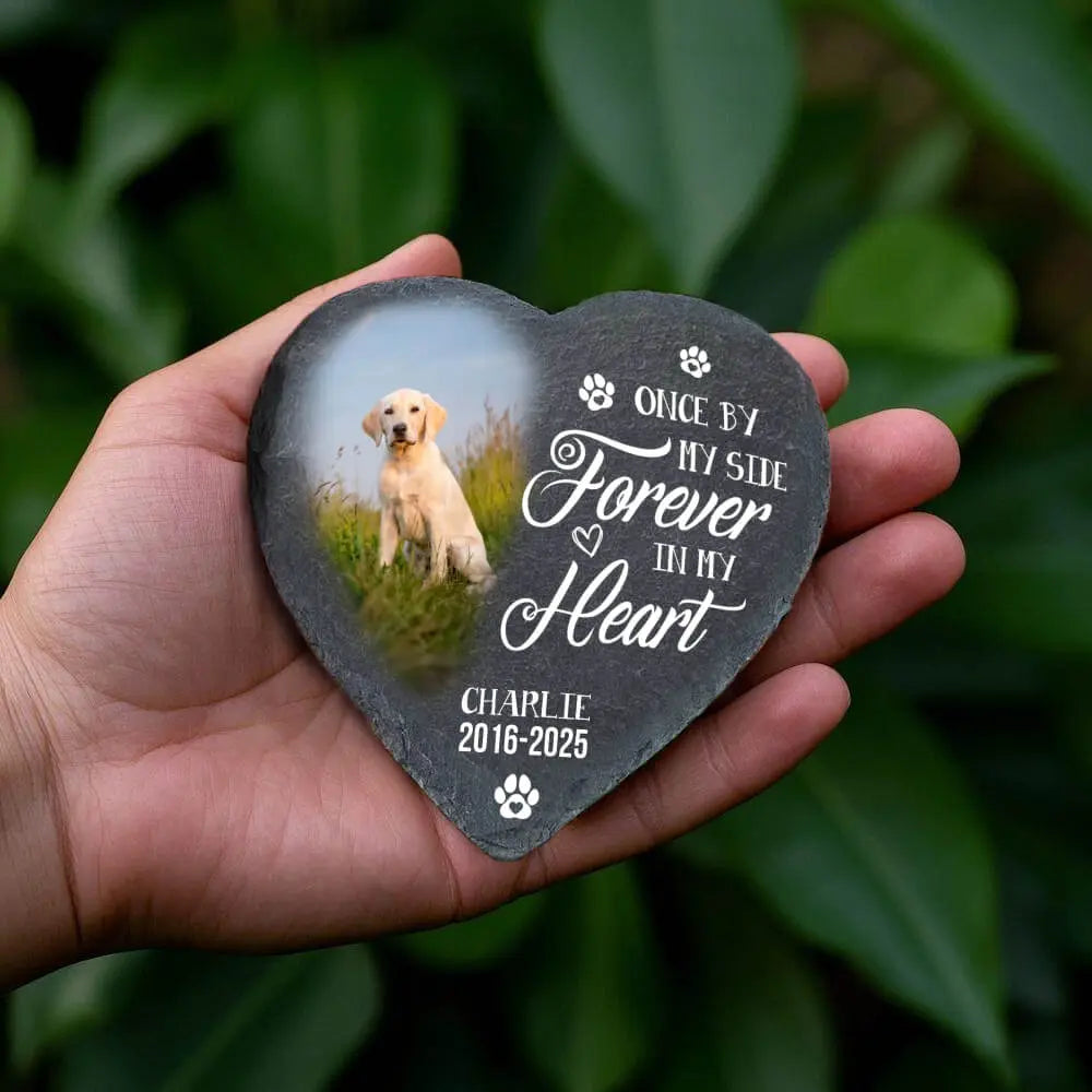 Heart-shaped pet memorial stone with a dog's photo and text, held in a hand with a green leafy background.